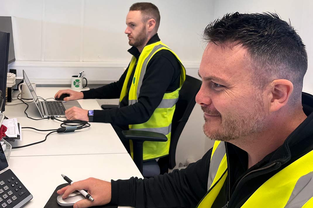 Two men in high-visibility vests working at a desk in an office.